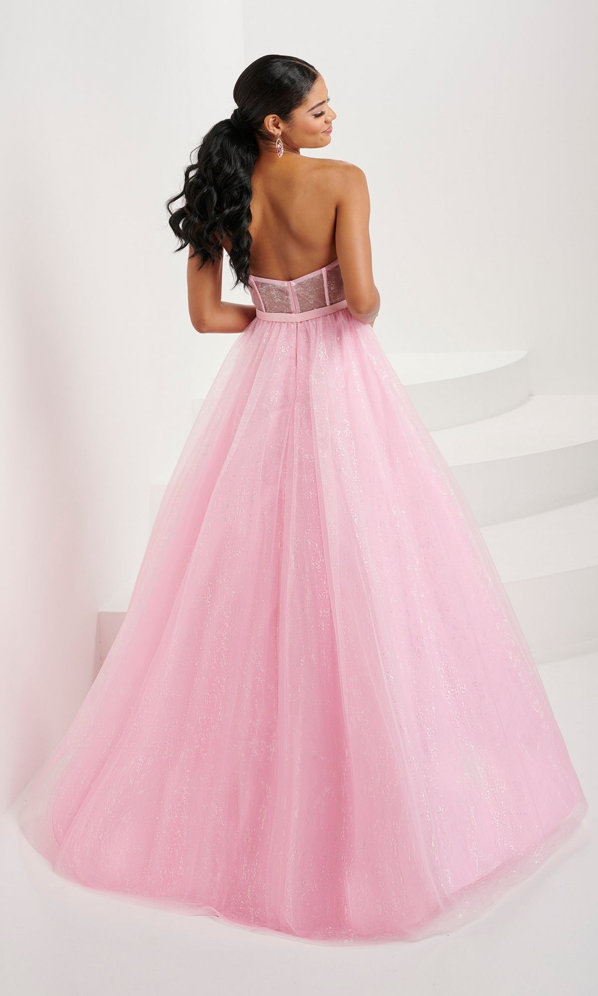 A woman with long, dark hair in a low ponytail models the Tiffany Strapless Glitter Pastel Prom Dress 16083, featuring a sparkling pink corset bodice, standing with her back to the camera against a white background.